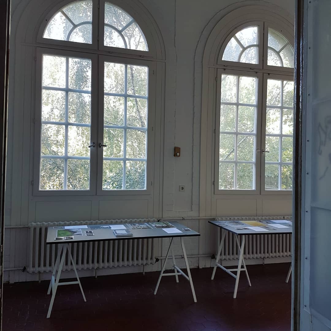 Ausstellungsansicht von zwei Tischen mit Bildern vor zwei Fenstern.
